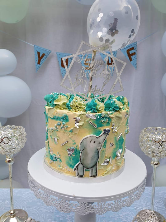 A eight inch vanilla strawberry buttercream cake, topped with a balloon, cake topper, textured buttercream, silver edible leaf, silver edible sprinkles and a hand painted elephant. 
